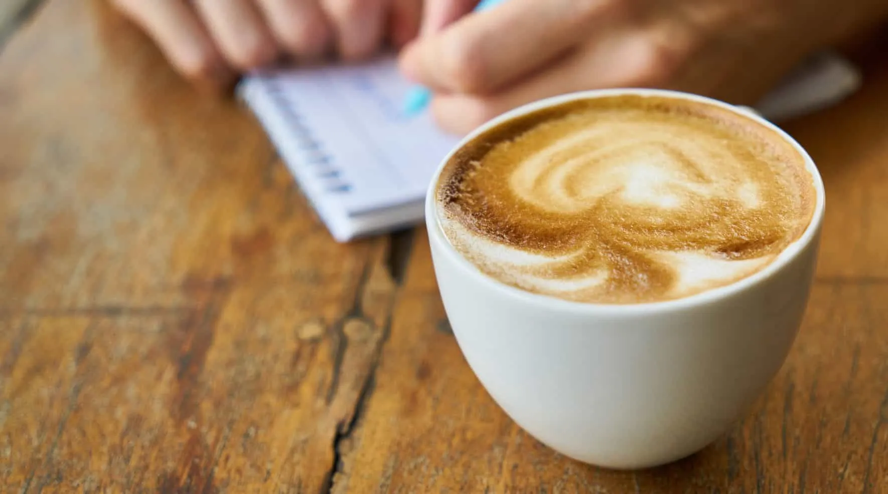 Tasse Cappuccino auf einem Holztisch, dahinter schreibt jemand in einen Notizblock – Java 15 Features