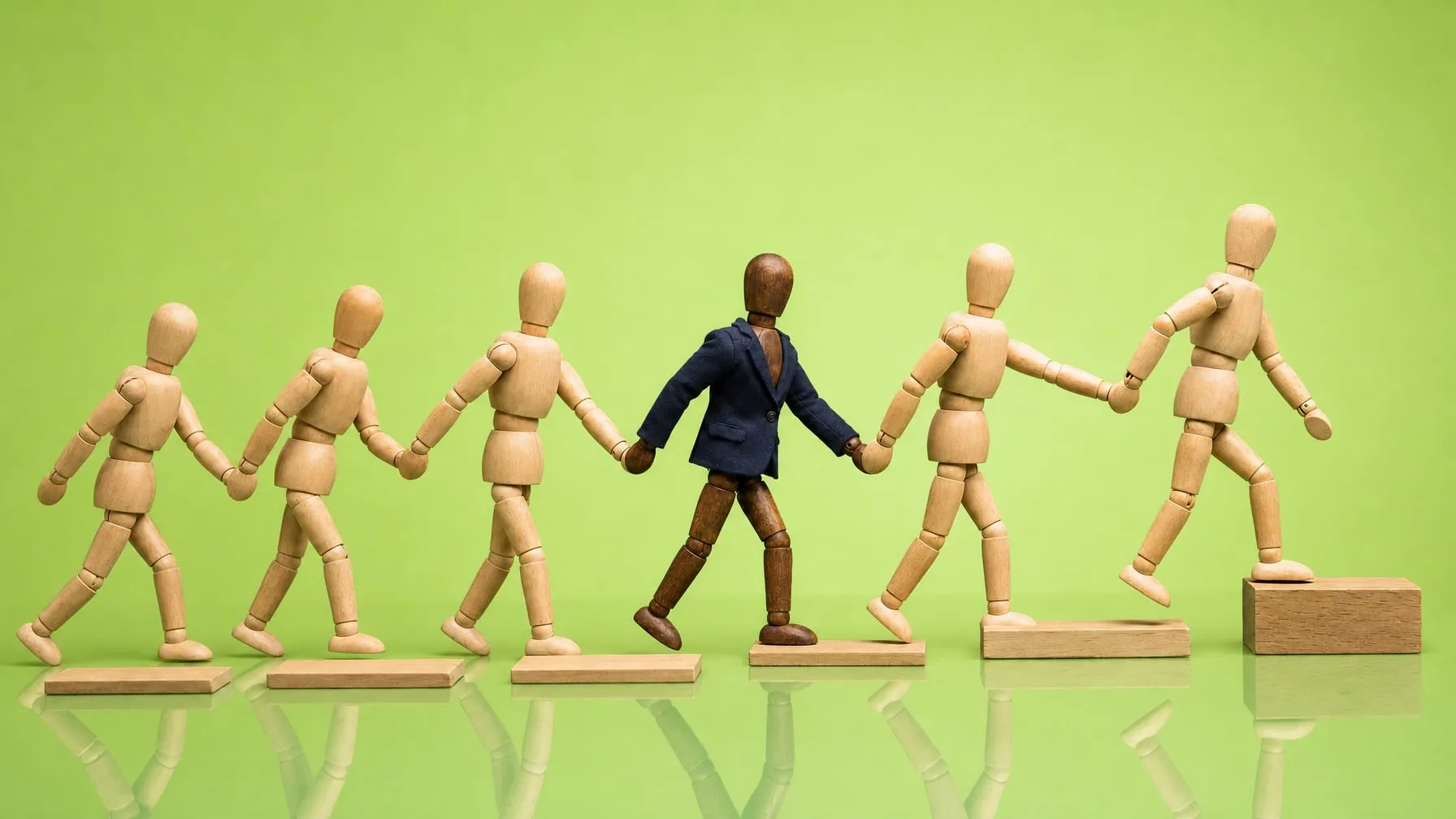 Sechs Holzgliederpuppen gehen Hand in Hand von links nach rechts über flache Holzstufen, die vorderste steigt auf die höchste, in der Mitte geht eine Puppe im Sakko mit