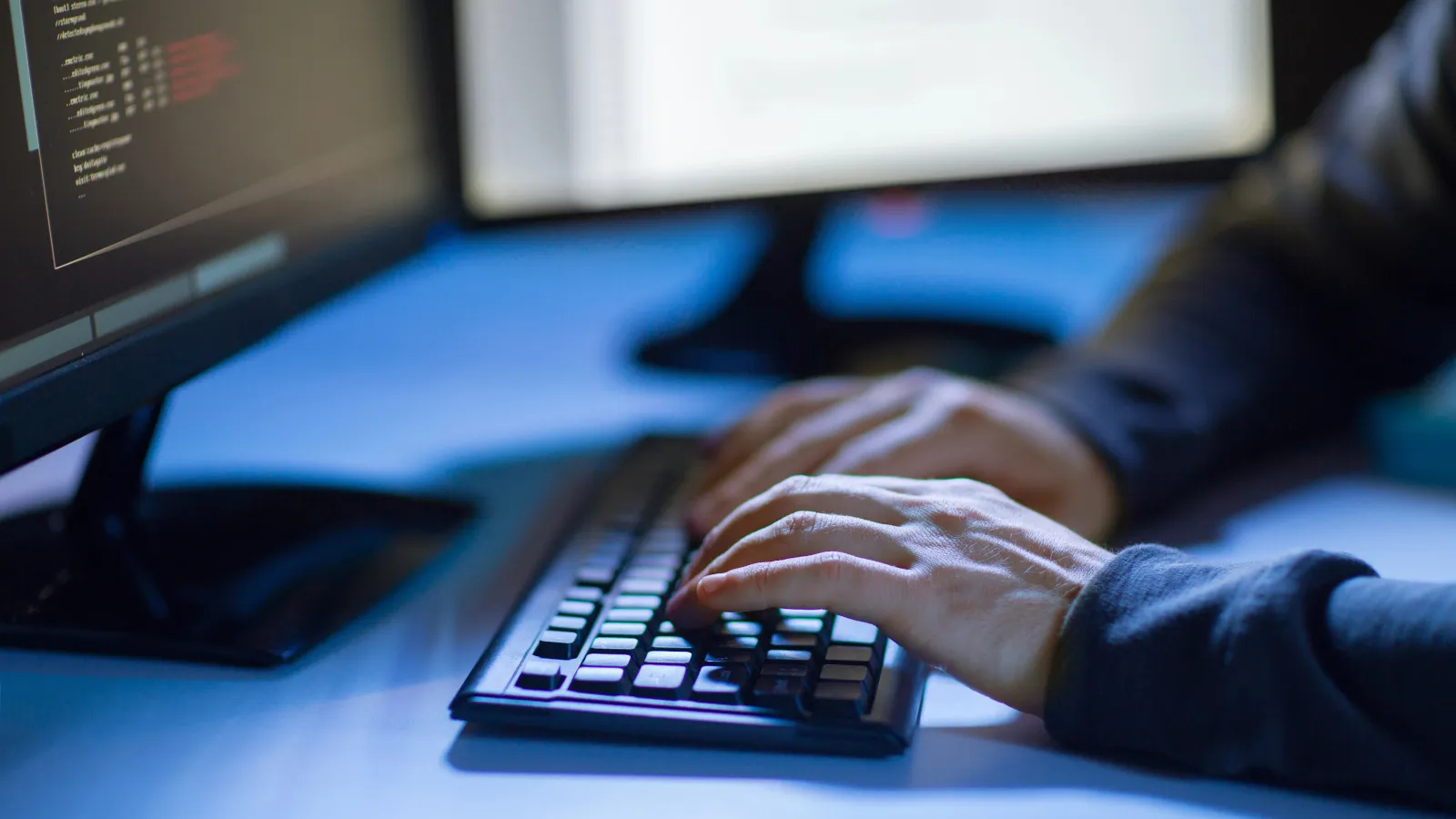 Hands typing on a keyboard in front of two monitors showing code